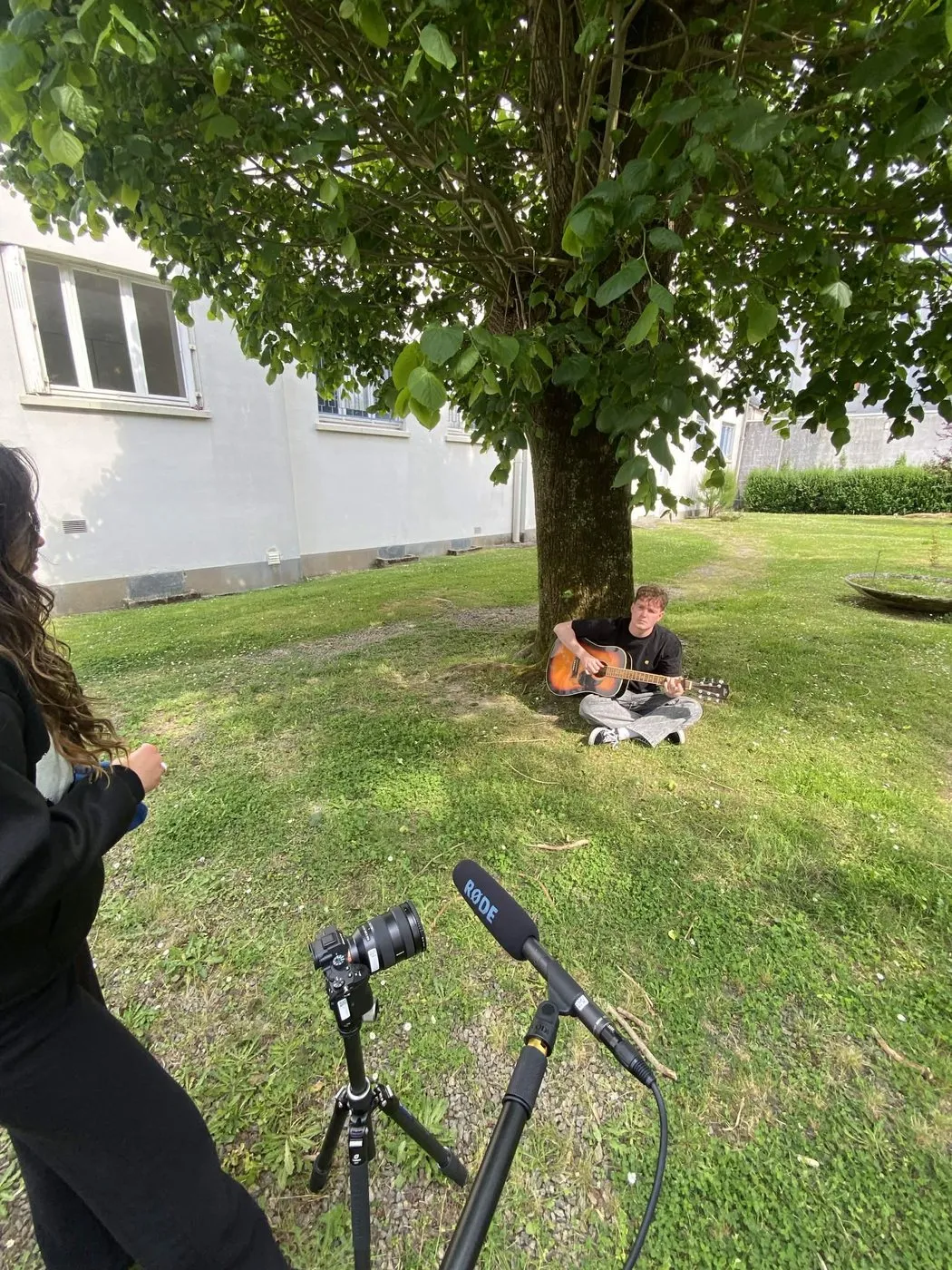 Un comédien joue de la guitare au pied d'un arbre, caméra sur trépied au premier plan
